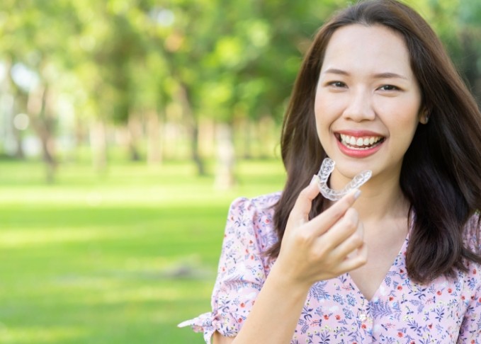 Smiling woman in a park holding an Invisalign aligner